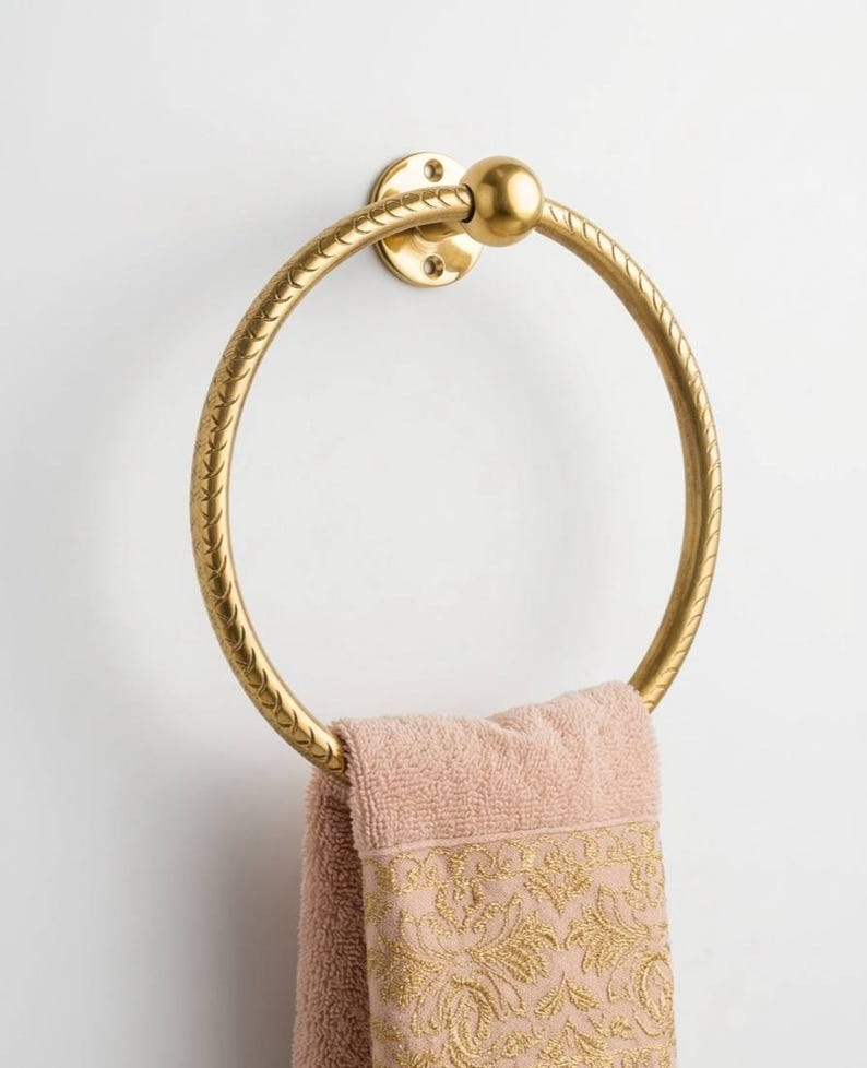 May include: A gold-colored towel ring with a textured design, mounted on a white wall. A pink towel with gold embroidery hangs from the ring, adding a touch of elegance to the bathroom decor.