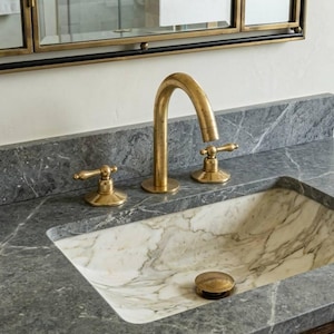 May include: A bathroom sink with a marble basin and a grey countertop. The tap and handles are gold-coloured. A gold-framed mirror is in the background.