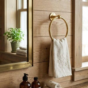 May include: A bathroom scene featuring a gold towel ring holding a folded beige towel with floral embroidery. The wall is made of light brown wood. There are rolled towels on a small plate and two brown bottles with labels.