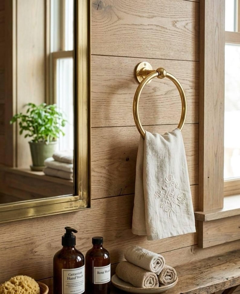 May include: A bathroom scene featuring a gold towel ring holding a folded beige towel with floral embroidery. The wall is made of light brown wood. There are rolled towels on a small plate and two brown bottles with labels.