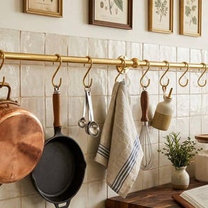May include: A kitchen with a brass pot rack holding kitchen utensils. A copper pan, cast iron frying pan, measuring spoons, a tea towel, and a whisk are suspended. Jars of flour and a cookbook are on the counter.