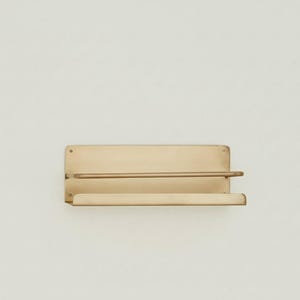 K&ouml;nnte beinhalten: Wandregal aus Messing. Es hat eine rechteckige R&uuml;ckplatte, eine horizontale Stange und ein unteres Regal. Das Messing hat einen warmen, goldenen Farbton. Das Design ist minimalistisch und modern.