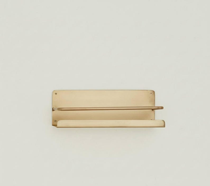 K&ouml;nnte beinhalten: Wandregal aus Messing. Es hat eine rechteckige R&uuml;ckplatte, eine horizontale Stange und ein unteres Regal. Das Messing hat einen warmen, goldenen Farbton. Das Design ist minimalistisch und modern.