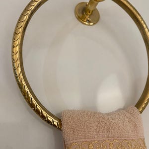 A gold-tone brass towel ring with a rope-like textured design hangs on a wall, A light pink hand towel with gold embroidered detailing hangs on the ring