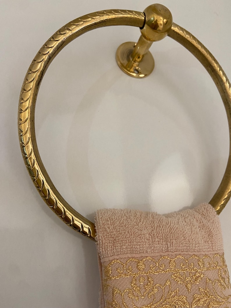 A gold-tone brass towel ring with a rope-like textured design hangs on a wall, A light pink hand towel with gold embroidered detailing hangs on the ring