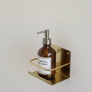 K&ouml;nnte beinhalten: Eine braune Glasflasche mit silbernem Pumpspender, beschriftet mit "BOTANICAL HAND SOAP", befindet sich in einem goldfarbenen, an der Wand befestigten Metallhalter. Der Halter hat eine rechteckige Basis und eine gebogene Halterung. Der Hintergrund ist eine schlichte, cremefarbene Wand.