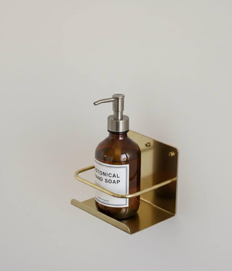 K&ouml;nnte beinhalten: Eine braune Glasflasche mit silbernem Pumpspender, beschriftet mit "BOTANICAL HAND SOAP", befindet sich in einem goldfarbenen, an der Wand befestigten Metallhalter. Der Halter hat eine rechteckige Basis und eine gebogene Halterung. Der Hintergrund ist eine schlichte, cremefarbene Wand.
