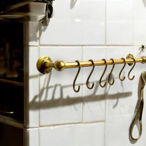 Könnte beinhalten: Ein Küchenutensilienhalter aus Messing mit sechs Haken, der an einer weißen gekachelten Wand montiert ist. Der Halter ist aus poliertem Messing gefertigt und hat ein abgerundetes Ende.