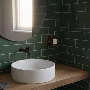 K&ouml;nnte beinhalten: Modernes Badezimmer mit einem wei&szlig;en, runden Waschbecken auf einer Holzplatte. Die W&auml;nde sind mit dunkelgr&uuml;nen rechteckigen Fliesen verkleidet. Ein schwarzer Wasserhahn und ein runder Spiegel sind an der Wand befestigt. Ein kleines Regal h&auml;lt einen Seifenspender.