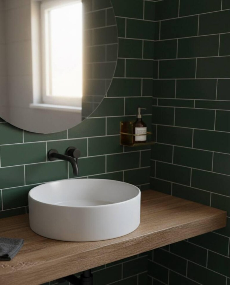K&ouml;nnte beinhalten: Modernes Badezimmer mit einem wei&szlig;en, runden Waschbecken auf einer Holzplatte. Die W&auml;nde sind mit dunkelgr&uuml;nen rechteckigen Fliesen verkleidet. Ein schwarzer Wasserhahn und ein runder Spiegel sind an der Wand befestigt. Ein kleines Regal h&auml;lt einen Seifenspender.