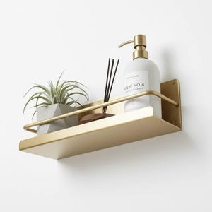 K&ouml;nnte beinhalten: Ein goldfarbenes Metallregal an einer wei&szlig;en Wand, das eine kleine Topfpflanze, eine Flasche Handseife, einen Holzdiffusor und Schilfst&auml;bchen enth&auml;lt. Auf der Handseifenflasche steht "SCENT NO. 4".