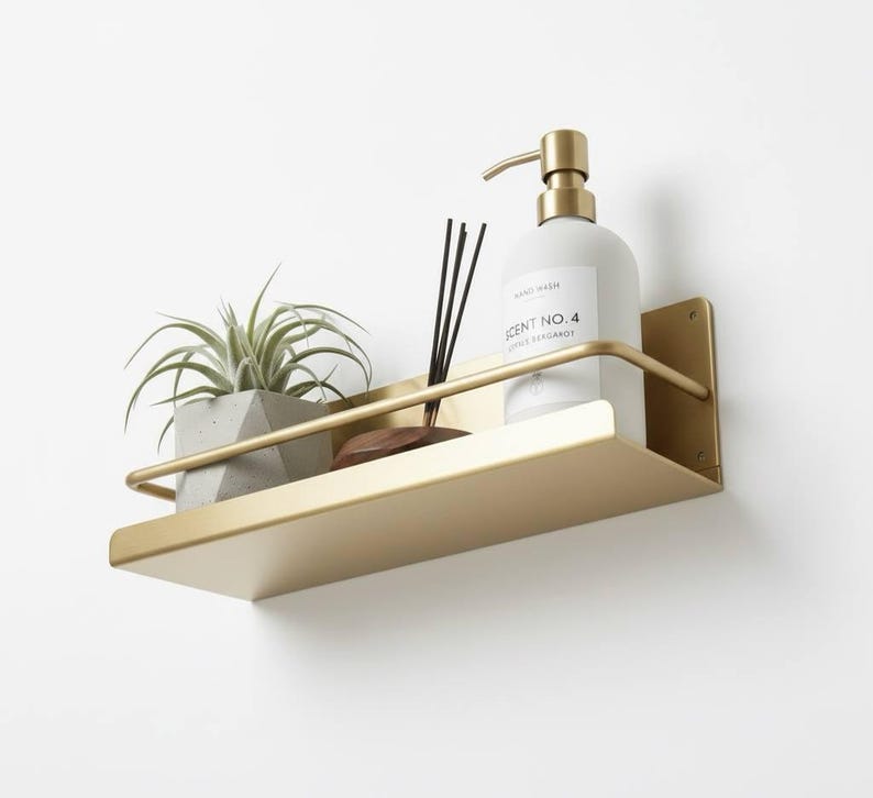 K&ouml;nnte beinhalten: Ein goldfarbenes Metallregal an einer wei&szlig;en Wand, das eine kleine Topfpflanze, eine Flasche Handseife, einen Holzdiffusor und Schilfst&auml;bchen enth&auml;lt. Auf der Handseifenflasche steht "SCENT NO. 4".
