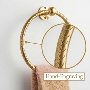 May include: A gold-colored towel ring with a decorative, hand-engraved design. The ring is circular and attached to a wall mount. A peach-colored towel with a gold floral pattern hangs from the ring. The text "Hand-Engraving" is visible.