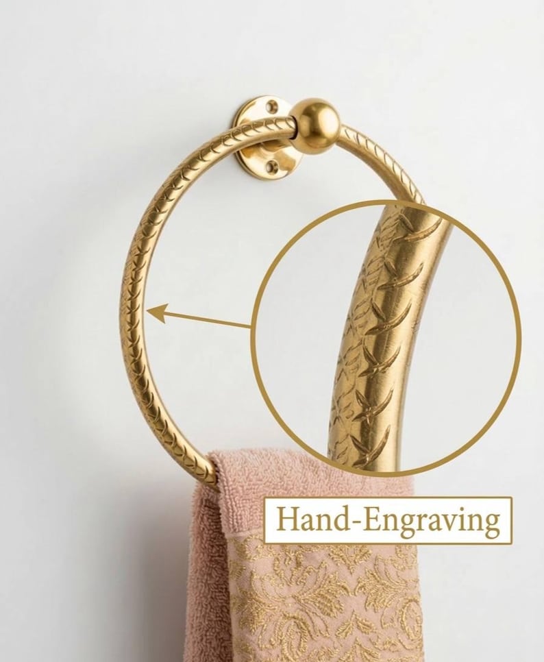 May include: A gold-colored towel ring with a decorative, hand-engraved design. The ring is circular and attached to a wall mount. A peach-colored towel with a gold floral pattern hangs from the ring. The text "Hand-Engraving" is visible.