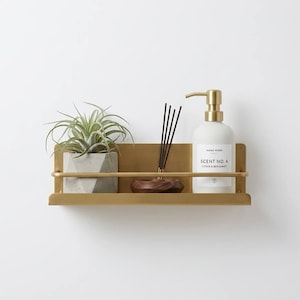 K&ouml;nnte beinhalten: Ein goldfarbenes Wandregal mit einer kleinen Topfpflanze, einer Holzschale mit R&auml;ucherst&auml;bchen und einer wei&szlig;en Handwaschflasche mit goldener Pumpe. Auf der Handwaschflasche steht "Scent No. 4 Citrus & Bergamot."