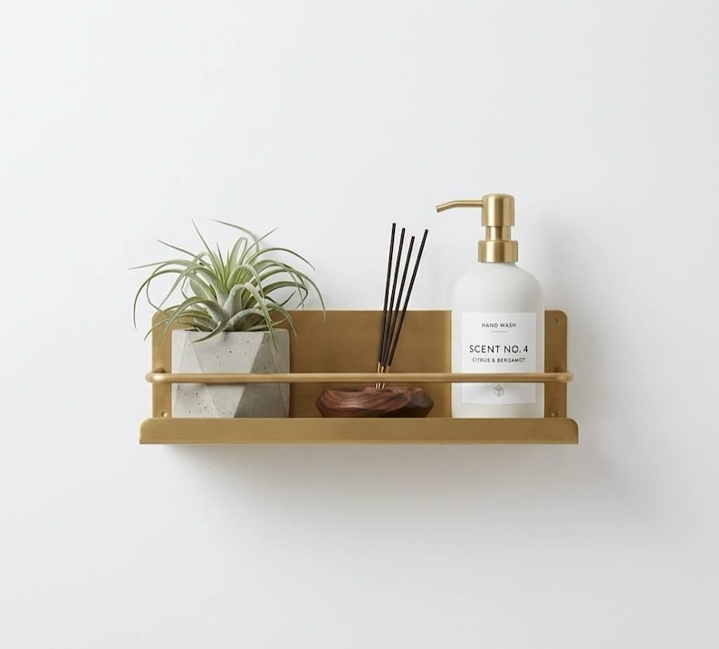 K&ouml;nnte beinhalten: Ein goldfarbenes Wandregal mit einer kleinen Topfpflanze, einer Holzschale mit R&auml;ucherst&auml;bchen und einer wei&szlig;en Handwaschflasche mit goldener Pumpe. Auf der Handwaschflasche steht "Scent No. 4 Citrus & Bergamot."