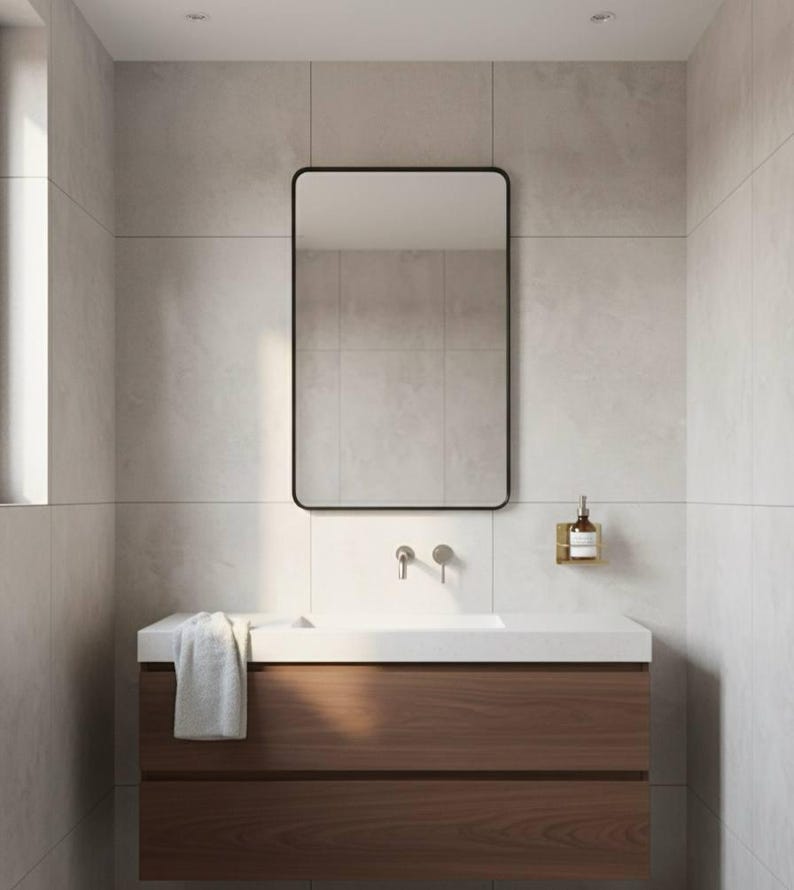 K&ouml;nnte beinhalten: Ein modernes Badezimmer mit einem rechteckigen, schwarz gerahmten Spiegel. Der Waschtisch hat eine wei&szlig;e Arbeitsplatte, einen Holzschrank mit zwei Schubladen und einen silbernen Wasserhahn. Ein Handtuch liegt auf der Theke, und ein Seifenspender ist an der Wand befestigt.