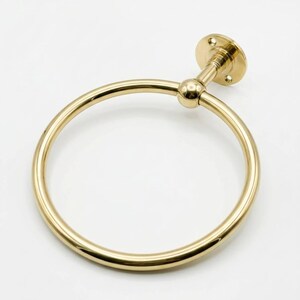 May include: A gold-colored towel ring with a circular design. The ring is attached to a wall mount with a small ball joint. The wall mount has a circular base with screw holes. The overall design is simple and elegant.