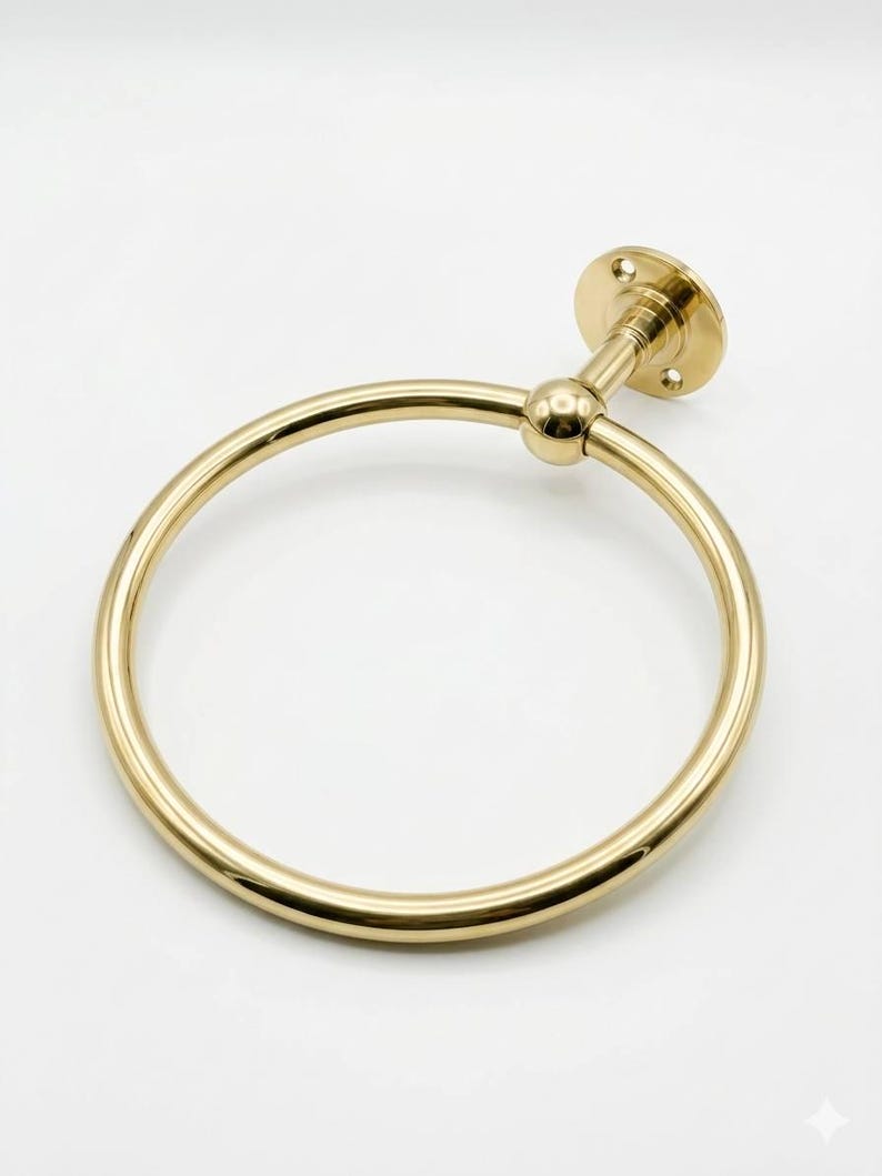 May include: A gold-colored towel ring with a circular design. The ring is attached to a wall mount with a small ball joint. The wall mount has a circular base with screw holes. The overall design is simple and elegant.