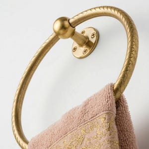 May include: A gold-colored towel ring with a textured design, mounted on a white wall. A pink towel with a gold embroidered border hangs from the ring. The ring is circular with a decorative pattern.