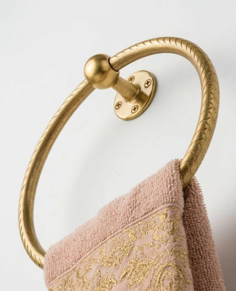 May include: A gold-colored towel ring with a textured design, mounted on a white wall. A pink towel with a gold embroidered border hangs from the ring. The ring is circular with a decorative pattern.