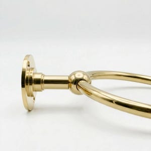 May include: A gold-colored towel ring with a circular design. The ring is attached to a round base with a connecting arm and a spherical detail. The metal has a polished finish, reflecting light.
