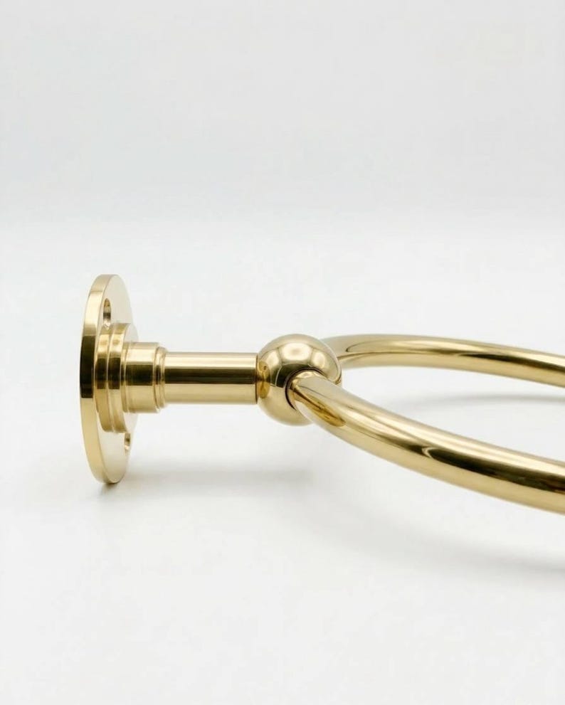 May include: A gold-colored towel ring with a circular design. The ring is attached to a round base with a connecting arm and a spherical detail. The metal has a polished finish, reflecting light.