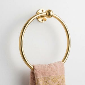 May include: A gold-colored towel ring mounted on a white wall. The ring is circular with a ball detail at the top. A pink towel with gold embroidery hangs from the ring, adding a touch of elegance to the bathroom.