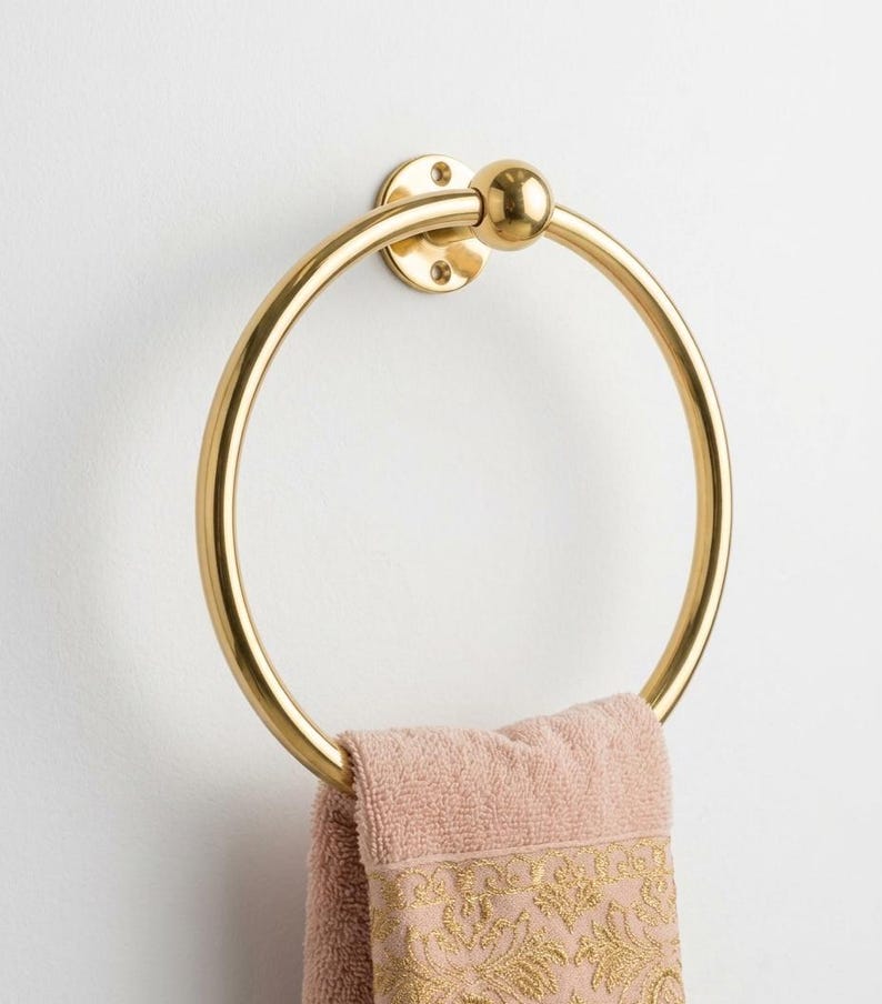May include: A gold-colored towel ring mounted on a white wall. The ring is circular with a ball detail at the top. A pink towel with gold embroidery hangs from the ring, adding a touch of elegance to the bathroom.
