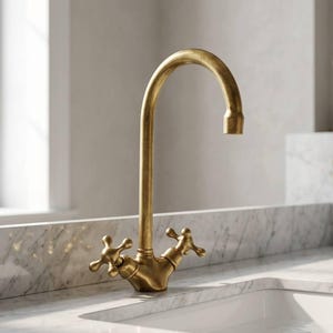 Può includere: Un rubinetto da cucina dorato con un beccuccio curvo e maniglie a forma di croce. Il rubinetto è montato su un piano di lavoro in marmo bianco. Il design è vintage ed elegante.