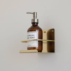 K&ouml;nnte beinhalten: Eine braune Glasflasche mit silbernem Pumpspender, beschriftet mit "BOTANICAL HAND SOAP", befindet sich in einem goldfarbenen, an der Wand befestigten Metallhalter. Der Halter hat eine rechteckige R&uuml;ckplatte und ein gebogenes Regal.