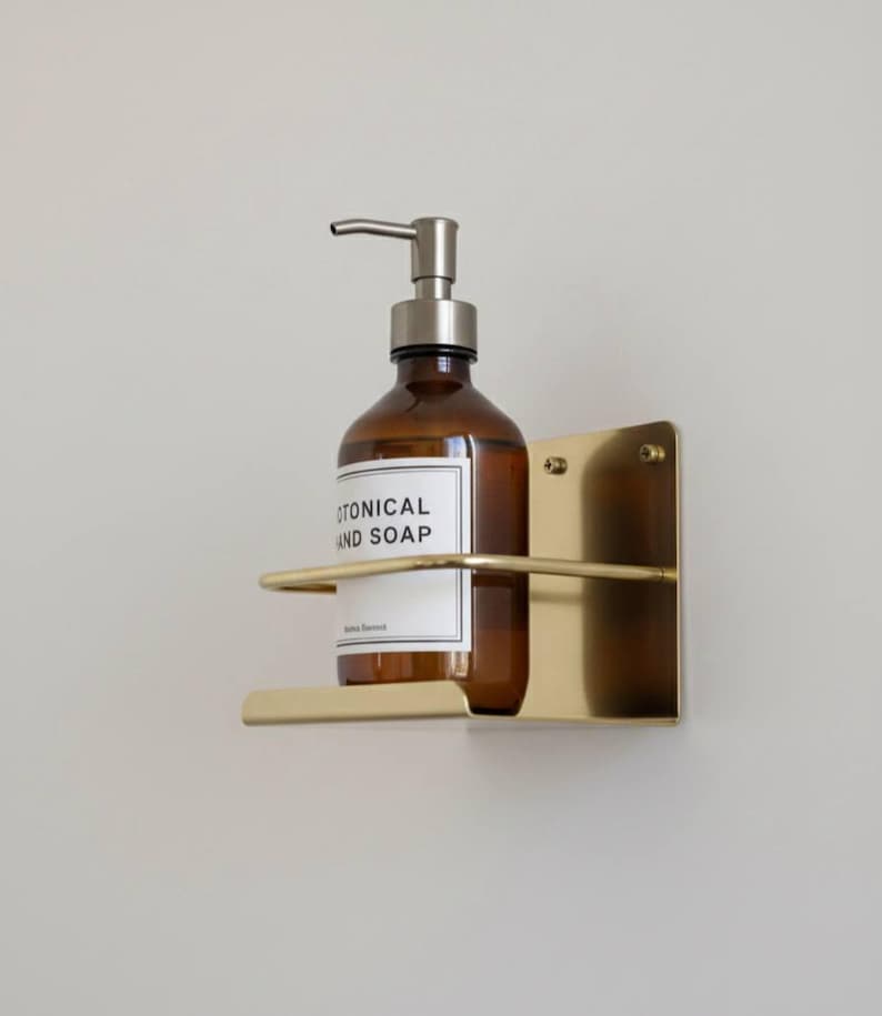 K&ouml;nnte beinhalten: Eine braune Glasflasche mit silbernem Pumpspender, beschriftet mit "BOTANICAL HAND SOAP", befindet sich in einem goldfarbenen, an der Wand befestigten Metallhalter. Der Halter hat eine rechteckige R&uuml;ckplatte und ein gebogenes Regal.