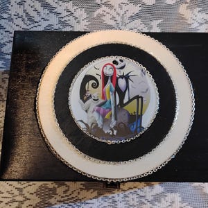 May include: A black wooden box with a circular design featuring characters from "The Nightmare Before Christmas." The design includes a white and silver border around a central image of Sally, Jack Skellington, and other characters.