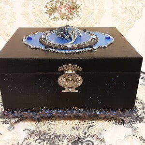 Op de afbeelding: Een zwarte houten doos met een blauw en zilveren decoratief deksel. Het deksel heeft een bloemmotief met strass steentjes en een zilveren ketting. De doos heeft een zilveren sluiting en een blauwe en zilveren rand rond de onderkant.