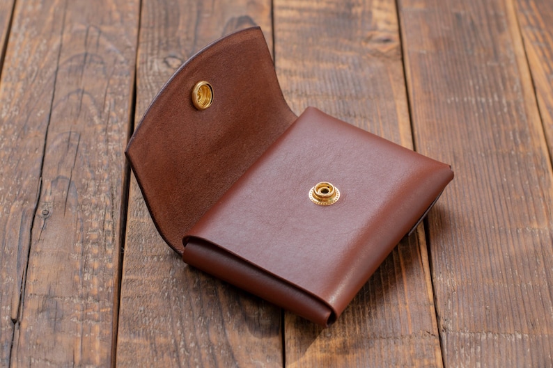 May include: A brown leather wallet with a fold-over flap and two brass snaps. The wallet is open and lying on a wooden surface.