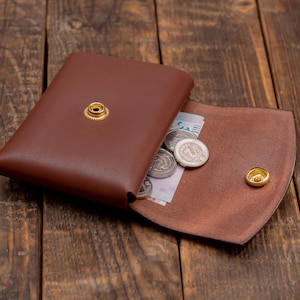 May include: A brown leather wallet with a flap closure and a snap button. The wallet is open and has coins and a bill inside.