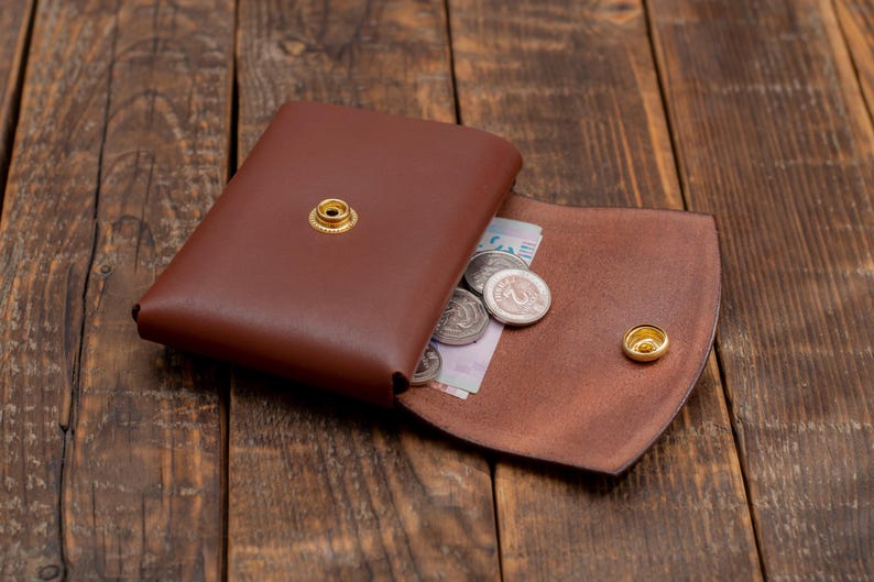 May include: A brown leather wallet with a flap closure and a snap button. The wallet is open and has coins and a bill inside.