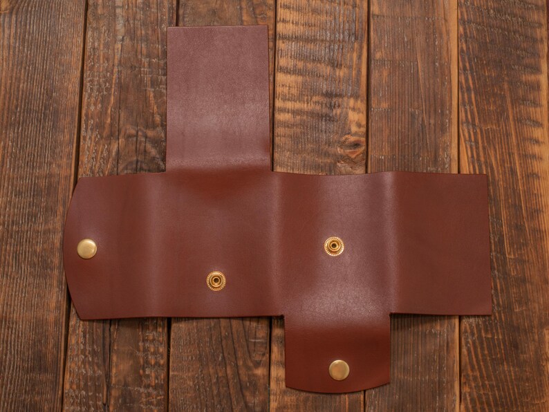 May include: A brown leather wallet with three folds and four brass snaps. The wallet is lying open on a wooden surface.