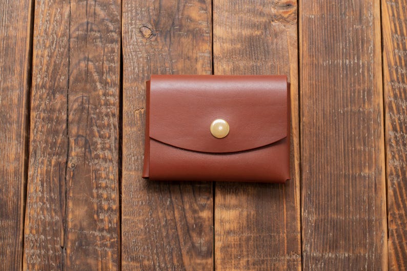 May include: A brown leather wallet with a brass button closure. The wallet is on a wooden surface.