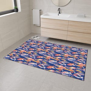Puede incluir: Una alfombra de baño rectangular con un patrón repetido de peces coloridos sobre un fondo azul oscuro. Los peces son en tonos naranja, amarillo, morado y blanco. La alfombra está en un baño.