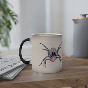 Può includere: Una tazza in ceramica bianca con manico nero, decorata con un'illustrazione di un ragno in bianco e nero. La tazza è una tazza che cambia colore quando si aggiunge liquido caldo.
