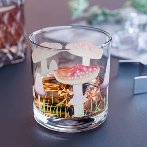 May include: A clear glass tumbler with a whimsical mushroom design. The mushrooms have red and white caps with white stems, set against a backdrop of green grass. The glass is filled with ice and a golden liquid.