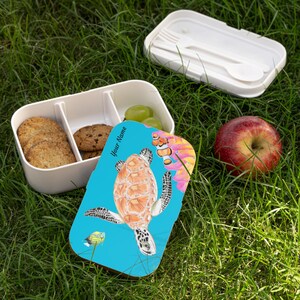 以下が含まれることがあります： 白い仕切り付きランチボックス。青い蓋にはウミガメのデザイン、クッキー、ブドウ、リンゴが緑の草の上に配置されています。蓋には「Your Name」の文字。別の容器にはフォークとスプーンが入っています。