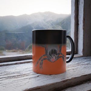 May include: A black and orange colour-changing mug with a detailed illustration of a spider on the orange side. The mug is sitting on a wooden surface.
