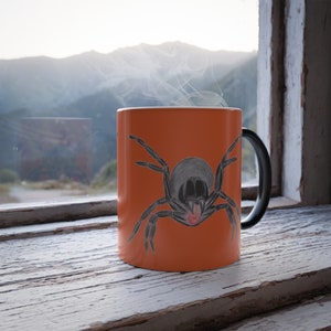 May include: Orange ceramic mug with a black handle. The mug features a black and white illustration of a spider on the front.