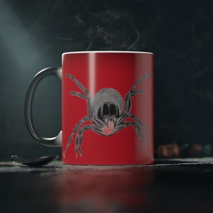 May include: A red ceramic mug featuring a black spider illustration. The spider has a black body and eight legs. The mug has a black handle.