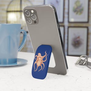May include: A smartphone is propped up by a blue phone grip with a watercolor octopus design. The octopus is light orange against the dark blue background. The phone is gray and has a silver trim.