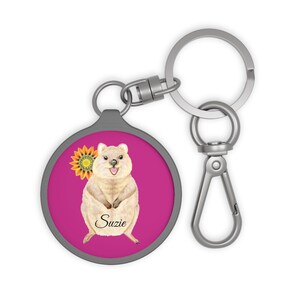 May include: A silver keychain with a round charm featuring a cartoon quokka with a sunflower behind it. The charm has a pink background and the name "Susie" written in black below the quokka.