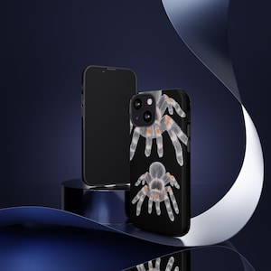 May include: Black smartphone case with a detailed illustration of a tarantula. The design shows the spider's body and legs in shades of grey and orange against a black background. Another black smartphone is also visible.