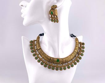 May include: A gold necklace and earrings set with green gemstone accents. The necklace is a crescent shape with intricate details and hangs from a black cord. The earrings are dangle style with a similar design.
